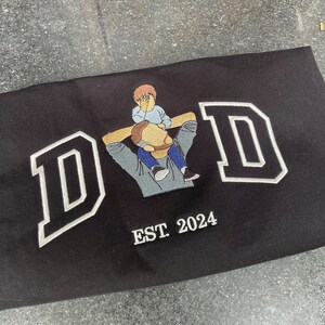 K&ouml;nnte beinhalten: Schwarzes besticktes T-Shirt mit dem Wort "DAD" in wei&szlig;en umrandeten Buchstaben. Ein kleines gesticktes Bild eines Vaters, der ein Kind auf seinen Schultern tr&auml;gt, ist mittig &uuml;ber dem Wort "DAD" platziert. Der Text "EST 2024" ist unter dem Wort "DAD" gestickt.