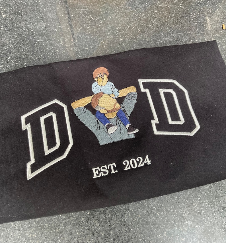 K&ouml;nnte beinhalten: Schwarzes besticktes T-Shirt mit dem Wort "DAD" in wei&szlig;en umrandeten Buchstaben. Ein kleines gesticktes Bild eines Vaters, der ein Kind auf seinen Schultern tr&auml;gt, ist mittig &uuml;ber dem Wort "DAD" platziert. Der Text "EST 2024" ist unter dem Wort "DAD" gestickt.