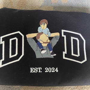 K&ouml;nnte beinhalten: Schwarzes T-Shirt mit Stickerei, mit einem wei&szlig;en Umriss des Wortes "DAD" und einem gestickten Bild von Vater und Sohn. Der Text "EST. 2024" ist unter dem Bild gestickt.