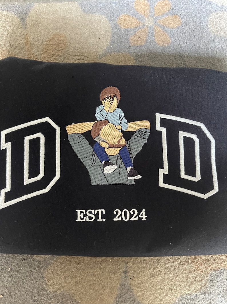 K&ouml;nnte beinhalten: Schwarzes T-Shirt mit Stickerei, mit einem wei&szlig;en Umriss des Wortes "DAD" und einem gestickten Bild von Vater und Sohn. Der Text "EST. 2024" ist unter dem Bild gestickt.