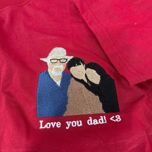 May include: A red t-shirt featuring an embroidered design of three figures. One figure wears a white hat and glasses, while the others are positioned beside them. Below the embroidery, the white text reads: Love you dad! <3