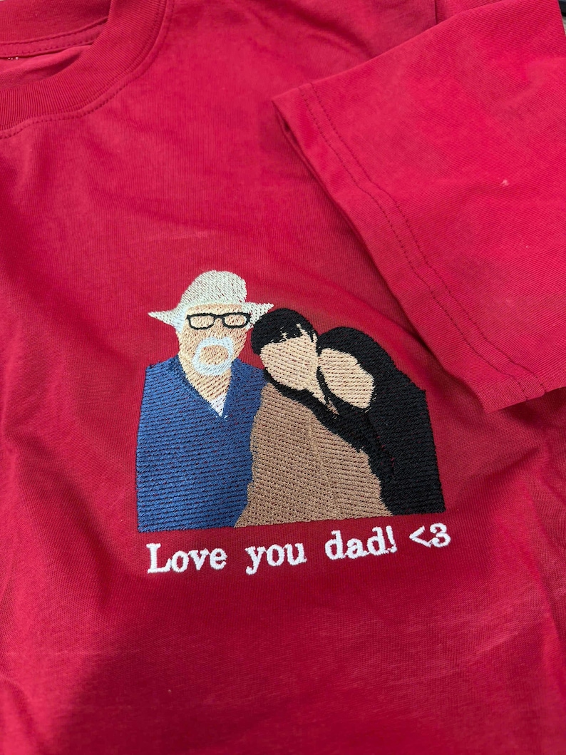 May include: A red t-shirt featuring an embroidered design of three figures. One figure wears a white hat and glasses, while the others are positioned beside them. Below the embroidery, the white text reads: Love you dad! <3