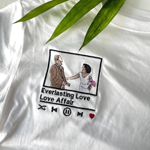 May include: A white t-shirt featuring an embroidered graphic of a couple in a square frame. Below the frame, the text reads Everlasting Love Love Affair, followed by music player icons including a shuffle, skip, pause, and a small red heart.