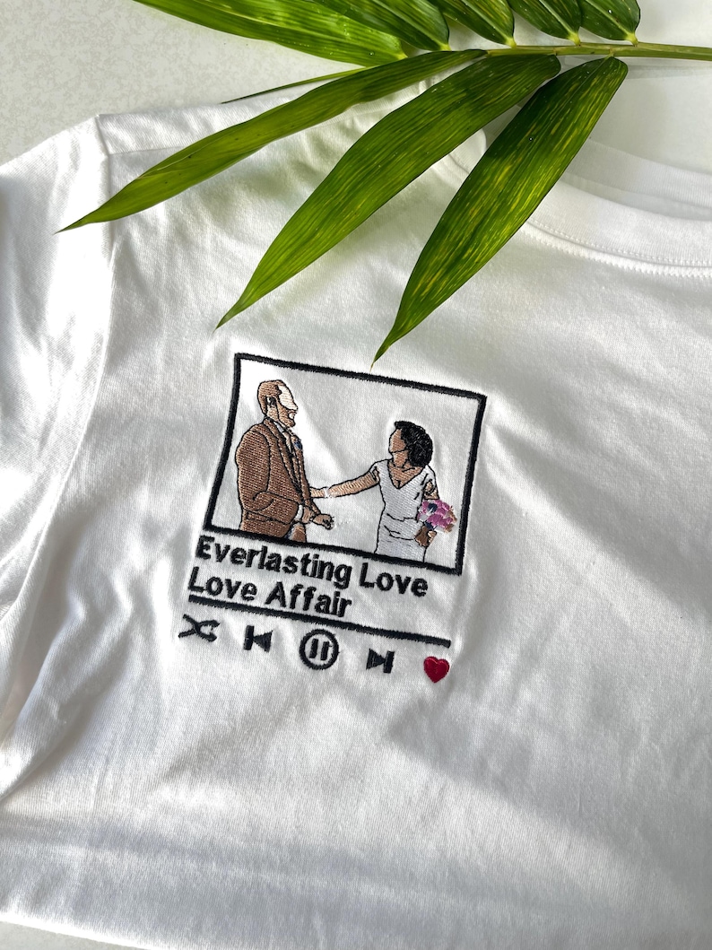 May include: A white t-shirt featuring an embroidered graphic of a couple in a square frame. Below the frame, the text reads Everlasting Love Love Affair, followed by music player icons including a shuffle, skip, pause, and a small red heart.