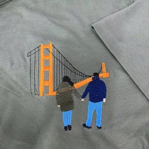 May include: An olive green t-shirt featuring an embroidered design of two figures holding hands while standing in front of an orange and black Golden Gate Bridge. The figures are wearing dark jackets and blue pants, rendered in detailed thread work.
