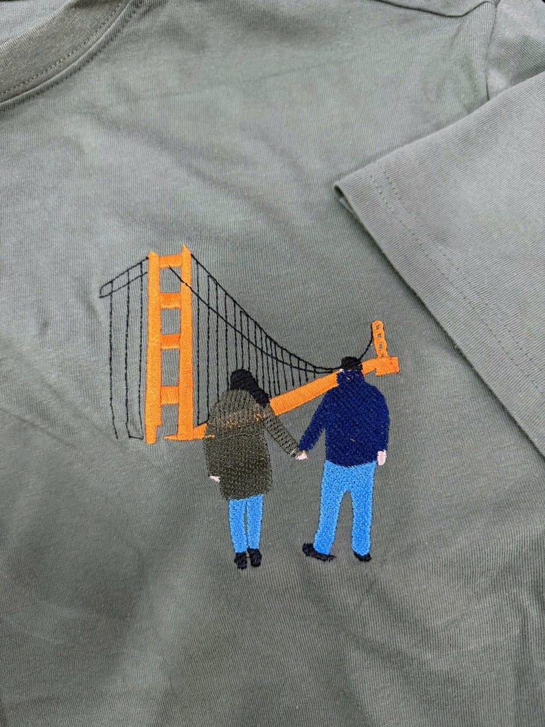 May include: An olive green t-shirt featuring an embroidered design of two figures holding hands while standing in front of an orange and black Golden Gate Bridge. The figures are wearing dark jackets and blue pants, rendered in detailed thread work.