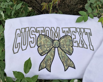 May include: White sweatshirt with the text "CUSTOM TEXT" in a bold, outlined font. A camouflage bow is centered between the words.