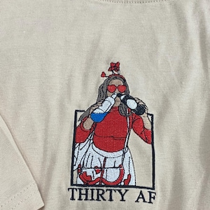 May include: A beige t-shirt featuring an embroidered graphic of a figure wearing heart-shaped sunglasses and a headband, drinking from two bottles. The figure wears a red top and white skirt, framed in a black box with the text THIRTY AF below.