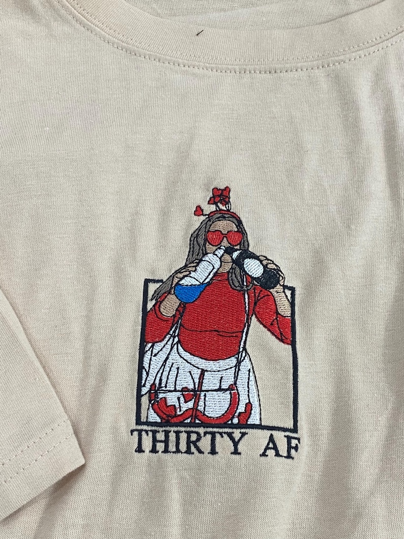 May include: A beige t-shirt featuring an embroidered graphic of a figure wearing heart-shaped sunglasses and a headband, drinking from two bottles. The figure wears a red top and white skirt, framed in a black box with the text THIRTY AF below.