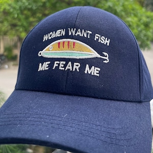 Könnte beinhalten: Eine marineblaue Baseballkappe mit weißem Sticktext, der "WOMEN WANT FISH" oben und "ME FEAR ME" unten zeigt, sowie einer zentralen Grafik eines gelben und hellgrünen Angelköders mit roten Streifen und einem kleinen Haken auf der rechten Seite.