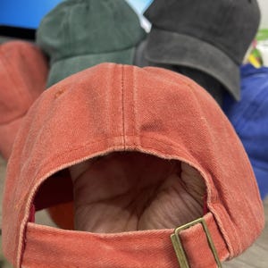 May include: A faded orange baseball cap with a curved brim and a fabric strap closure featuring a brass-toned metal slider buckle. The cap has a distressed, washed-out texture and visible stitching along the panels and the adjustable back strap.