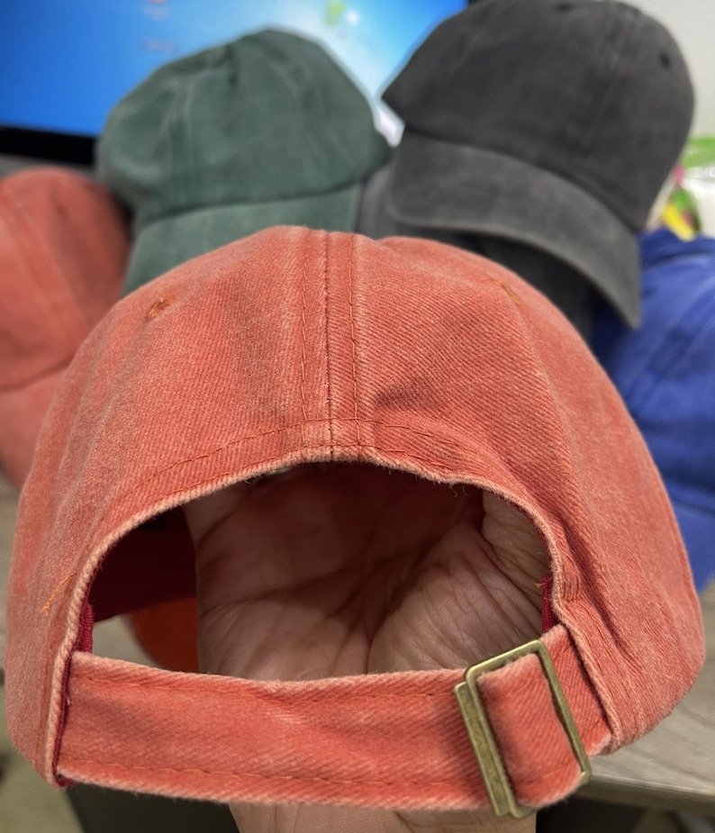 May include: A faded orange baseball cap with a curved brim and a fabric strap closure featuring a brass-toned metal slider buckle. The cap has a distressed, washed-out texture and visible stitching along the panels and the adjustable back strap.
