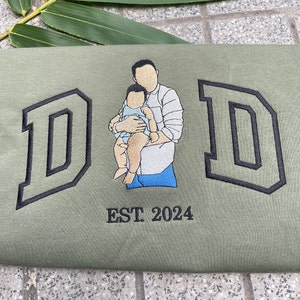 K&ouml;nnte beinhalten: Ein gr&uuml;nes T-Shirt mit schwarzen gestickten Buchstaben, die "DAD" buchstabieren, und einem Bild eines Vaters, der ein Baby h&auml;lt. Der Text "EST 2024" ist unter dem Bild gestickt.