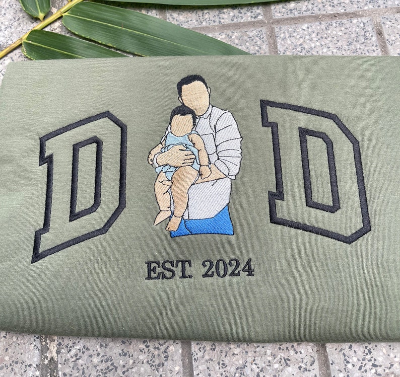 K&ouml;nnte beinhalten: Ein gr&uuml;nes T-Shirt mit schwarzen gestickten Buchstaben, die "DAD" buchstabieren, und einem Bild eines Vaters, der ein Baby h&auml;lt. Der Text "EST 2024" ist unter dem Bild gestickt.