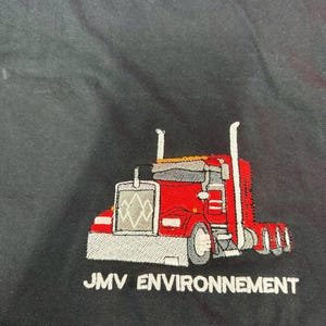 May include: A black t-shirt featuring an embroidered graphic of a red semi-truck with a silver grille and exhaust stacks. Below the truck, the text JMV ENVIRONNEMENT is embroidered in white capital letters against the black fabric background.