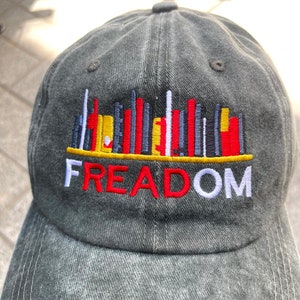 May include: A gray denim baseball cap with a colorful graphic of a city skyline and the word "FREEDOM" embroidered in red, white, and yellow.