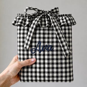 May include: A rectangular fabric pouch featuring a black and white gingham check pattern. The top edge has a ruffled trim and a tied fabric bow. The name Ava is embroidered in dark blue script lettering on the front center of the pouch.