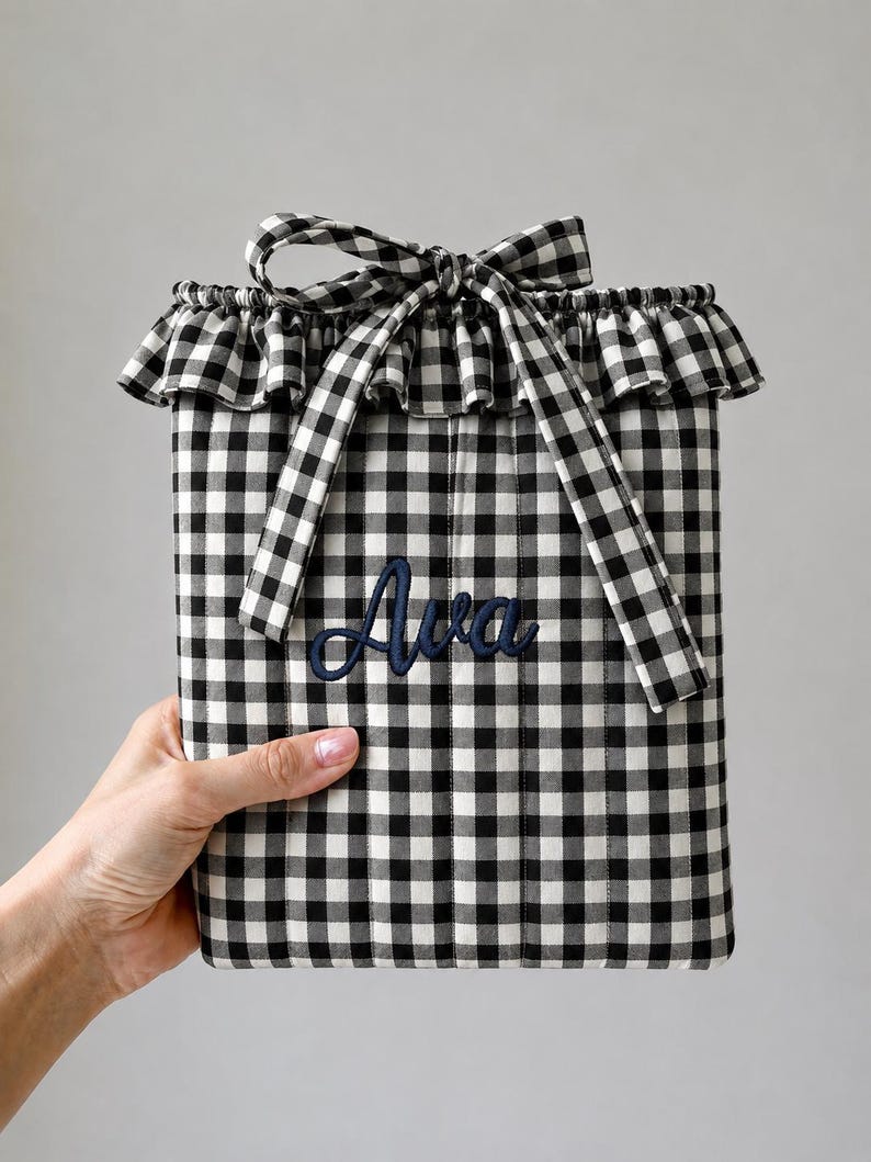 May include: A rectangular fabric pouch featuring a black and white gingham check pattern. The top edge has a ruffled trim and a tied fabric bow. The name Ava is embroidered in dark blue script lettering on the front center of the pouch.
