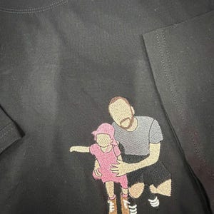 May include: A black t-shirt featuring an embroidered design of a person kneeling while holding a small child. The child wears a pink dress and hat, while the adult wears a gray t-shirt and black shorts. The figures have no facial features.