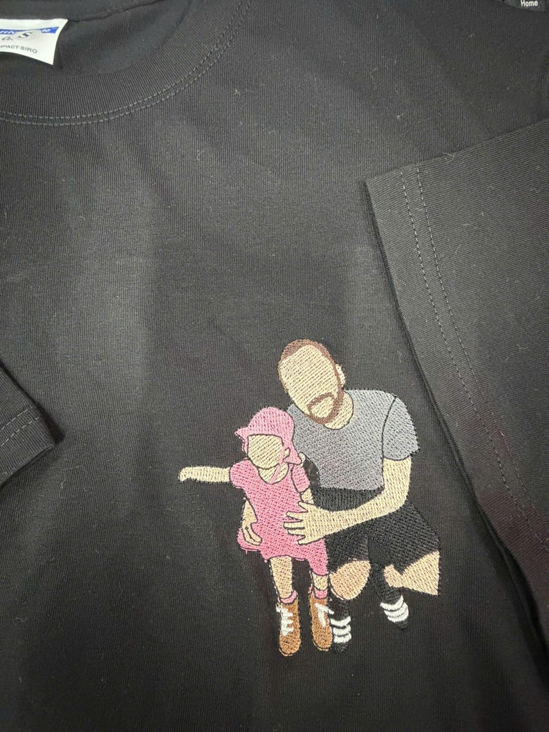 May include: A black t-shirt featuring an embroidered design of a person kneeling while holding a small child. The child wears a pink dress and hat, while the adult wears a gray t-shirt and black shorts. The figures have no facial features.