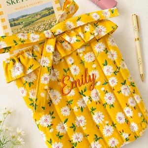 May include: A quilted yellow fabric sleeve featuring a white daisy print with green leaves, a ruffled top edge, and a tie closure. The name Emily is embroidered in orange script on the front. A book titled Sunday Sketching, a pink lipstick, and a pen are nearby.