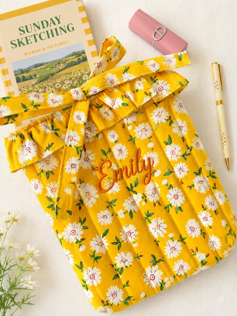 May include: A quilted yellow fabric sleeve featuring a white daisy print with green leaves, a ruffled top edge, and a tie closure. The name Emily is embroidered in orange script on the front. A book titled Sunday Sketching, a pink lipstick, and a pen are nearby.