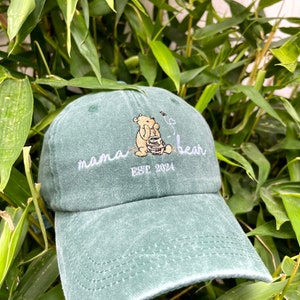 May include: A green, distressed, baseball cap with an embroidered design of a bear sitting in a honey pot. The text "mama bear est. 2024" is embroidered on the front of the cap.