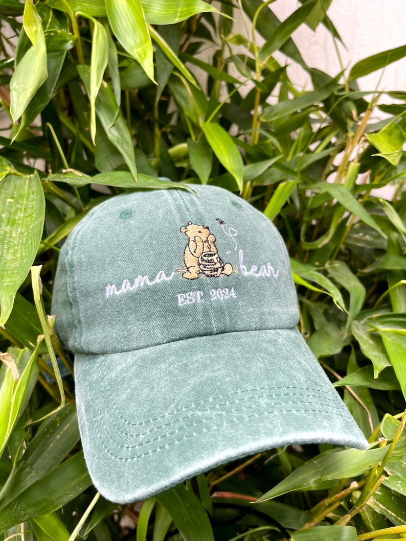 May include: A green, distressed, baseball cap with an embroidered design of a bear sitting in a honey pot. The text "mama bear est. 2024" is embroidered on the front of the cap.