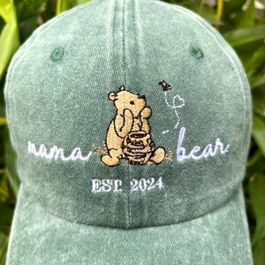 May include: Green baseball cap with embroidered Winnie the Pooh character holding a honey pot, with the text "mama bear" and "EST 2024" in white embroidery.