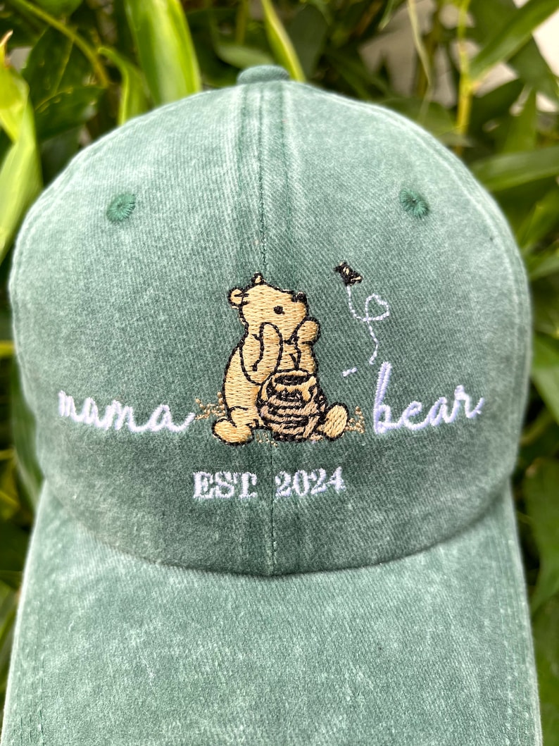May include: Green baseball cap with embroidered Winnie the Pooh character holding a honey pot, with the text "mama bear" and "EST 2024" in white embroidery.