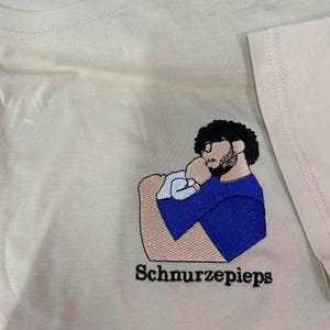 May include: A cream-colored t-shirt featuring an embroidered graphic of a person with dark curly hair, glasses, and a beard holding a baby. The person wears a blue shirt. The text "Schnurzepieps" is embroidered in black below the graphic.