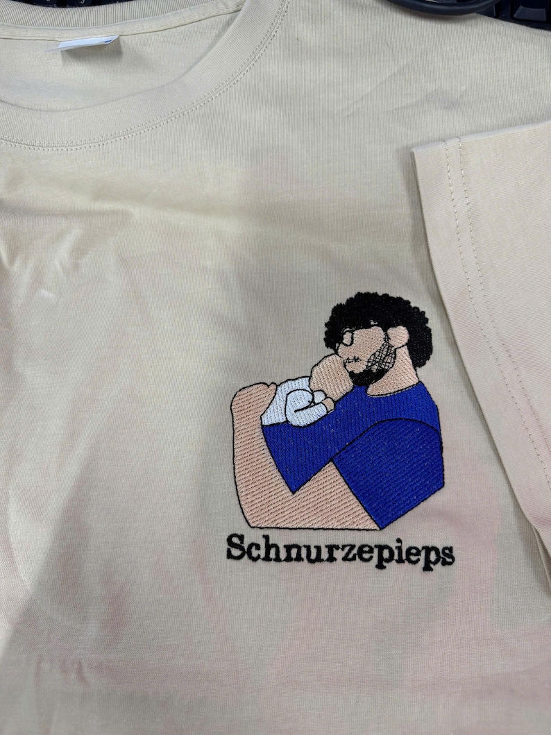 May include: A cream-colored t-shirt featuring an embroidered graphic of a person with dark curly hair, glasses, and a beard holding a baby. The person wears a blue shirt. The text "Schnurzepieps" is embroidered in black below the graphic.