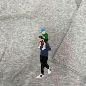 May include: A heather gray t-shirt featuring an embroidered design of a person with a beard wearing a dark blue long-sleeve shirt and black pants, carrying a small child on their shoulders who wears a green shirt and a bright blue baseball cap.
