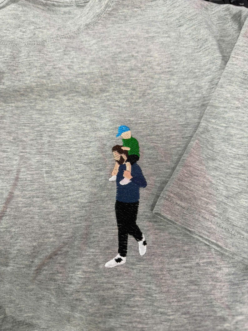 May include: A heather gray t-shirt featuring an embroidered design of a person with a beard wearing a dark blue long-sleeve shirt and black pants, carrying a small child on their shoulders who wears a green shirt and a bright blue baseball cap.