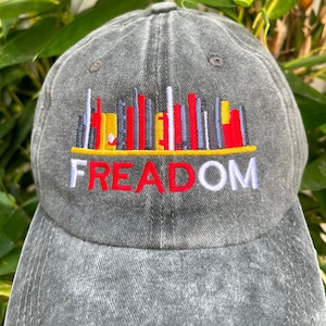 May include: A gray denim baseball cap with an embroidered design of a cityscape with the word "FREEDOM" underneath. The cityscape is made up of red, yellow, white, and black vertical lines.