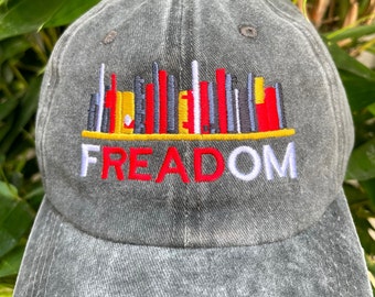May include: A gray denim baseball cap with an embroidered design of a cityscape with the word "FREEDOM" underneath. The cityscape is made up of red, yellow, white, and black vertical lines.