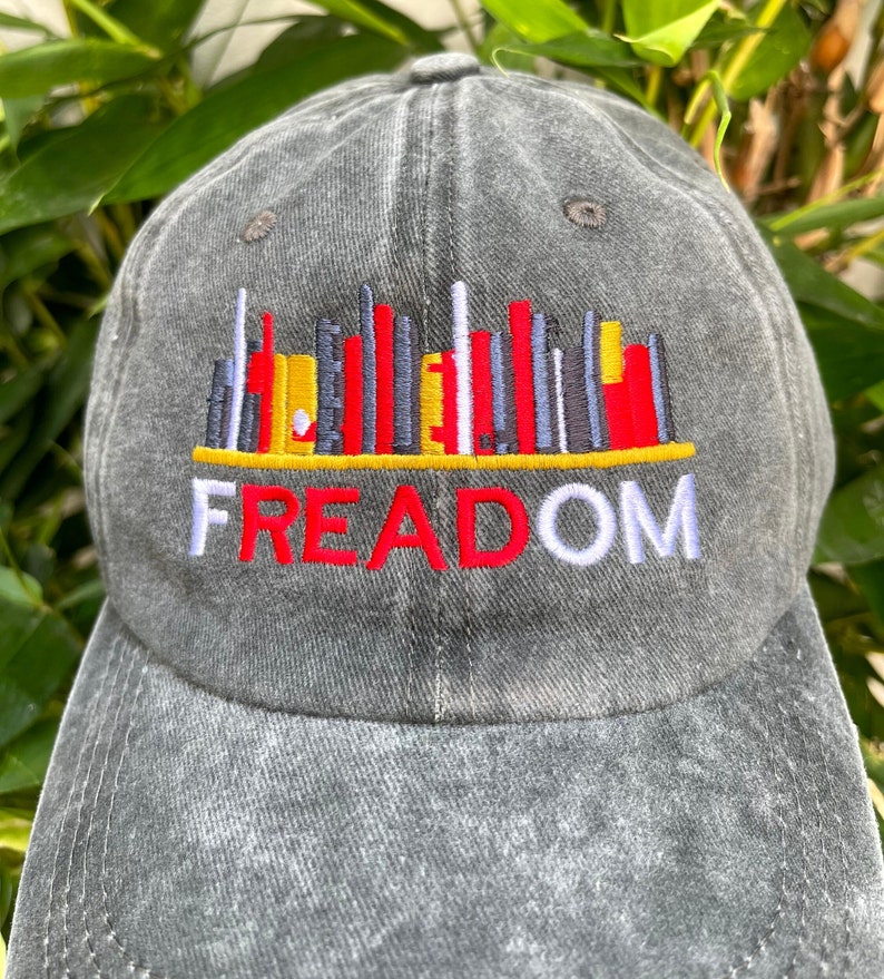 May include: A gray denim baseball cap with an embroidered design of a cityscape with the word "FREEDOM" underneath. The cityscape is made up of red, yellow, white, and black vertical lines.