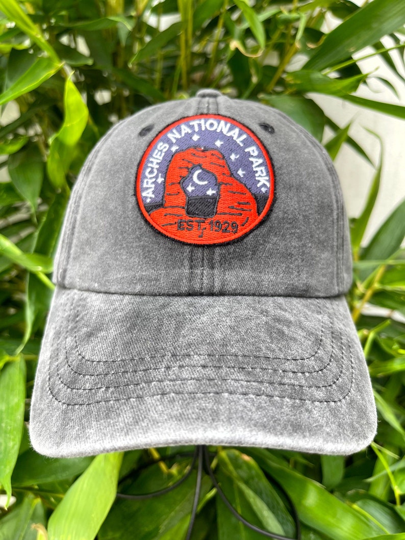 May include: A black, distressed, cotton baseball cap with an embroidered patch featuring the Arches National Park logo. The logo is a red circle with a white outline and the text "Arches National Park" in white. Inside the circle is a stylized image of Delicate Arch with a crescent moon and stars. The text "Est. 1929" is below the arch.