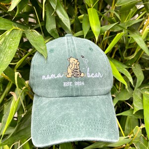 May include: A green, distressed, baseball cap with an embroidered image of a bear holding a honey pot. The text "mama bear" and "EST 2024" is also embroidered on the cap.