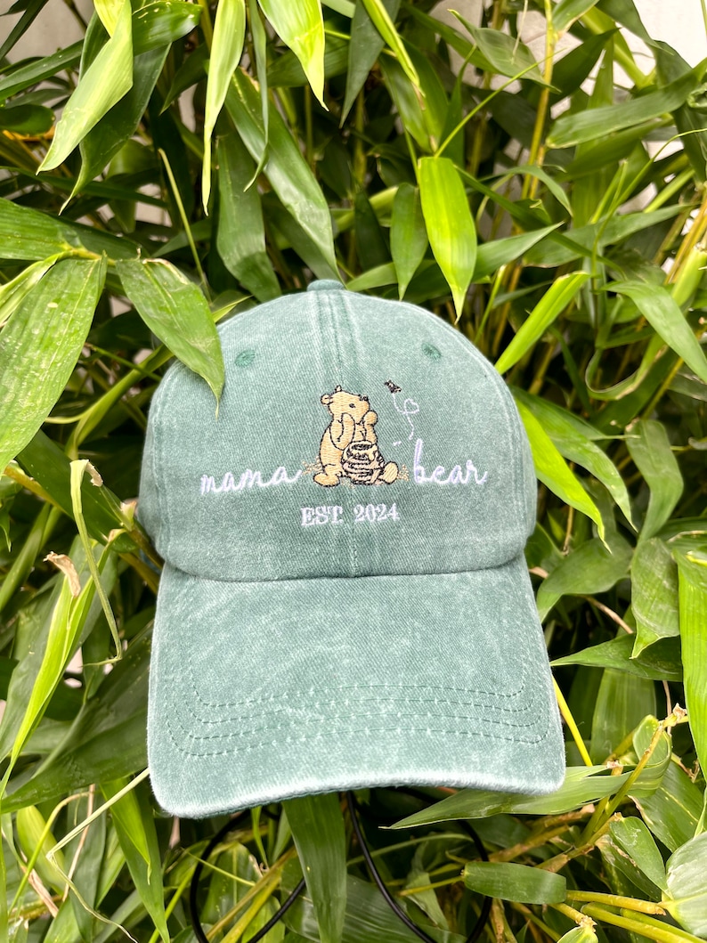 May include: A green, distressed, baseball cap with an embroidered image of a bear holding a honey pot. The text "mama bear" and "EST 2024" is also embroidered on the cap.