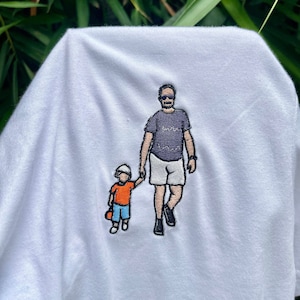 May include: A white t-shirt featuring an embroidered graphic of an adult and a small child walking while holding hands. The adult wears a dark purple t-shirt, white shorts, and sunglasses. The child wears an orange shirt, blue shorts, and a white hat.