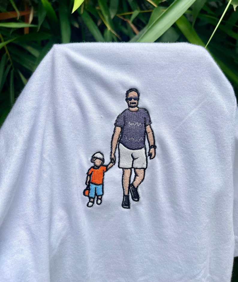 May include: A white t-shirt featuring an embroidered graphic of an adult and a small child walking while holding hands. The adult wears a dark purple t-shirt, white shorts, and sunglasses. The child wears an orange shirt, blue shorts, and a white hat.