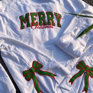 Könnte beinhalten: Weißer Langarm-Pullover mit grünen und roten Glitzerverzierungen. Die Rückseite des Pullovers trägt die Aufschrift "Merry Christmas" in grünem und rotem Glitzer. Die Ärmel haben grüne Glitzerbögen.