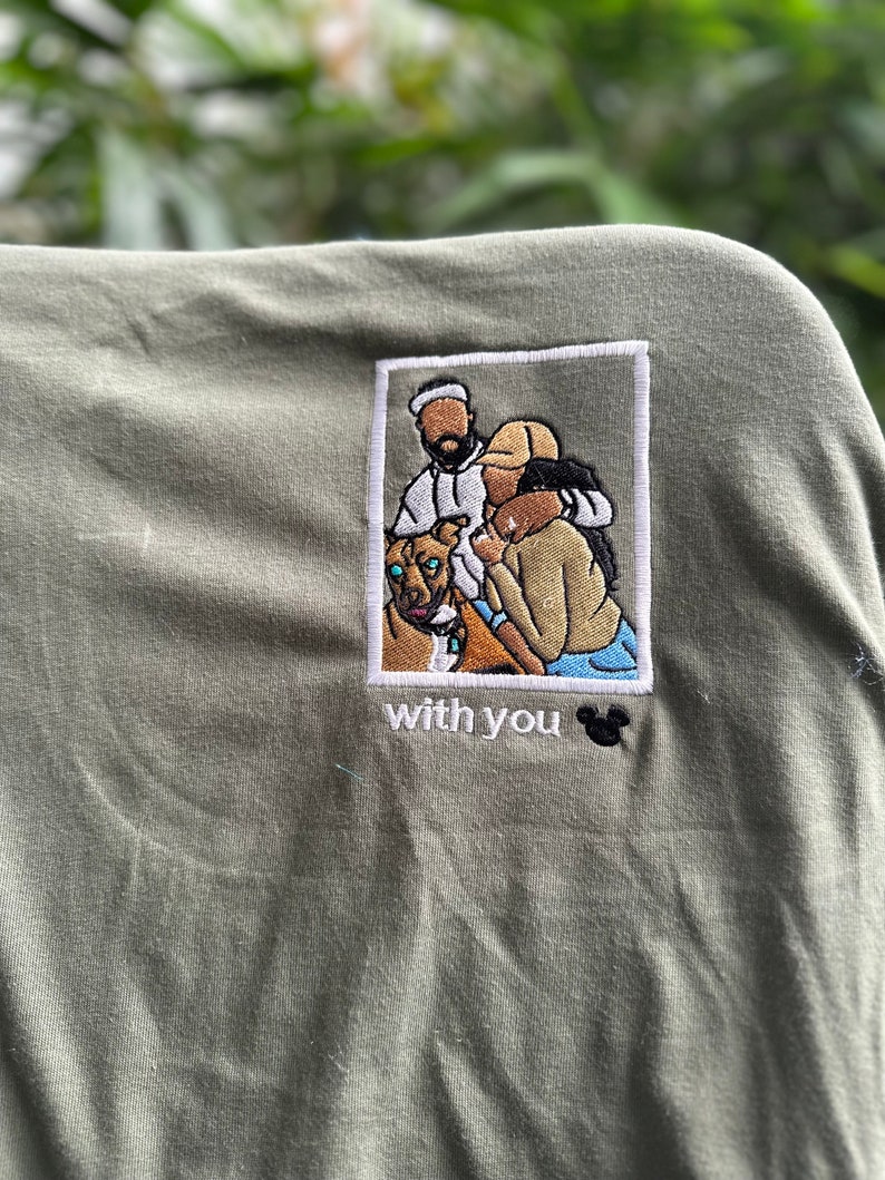 May include: An olive green t-shirt featuring a rectangular embroidered patch. The patch depicts a person in a white hoodie, a person in a tan shirt, and a brown dog. Below the patch, the text "with you" is embroidered with a black mouse icon.