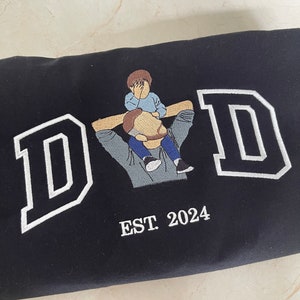 K&ouml;nnte beinhalten: Schwarzer Sweatshirt mit wei&szlig; gesticktem Text "DAD" und einem kleinen gestickten Bild von Vater und Sohn. Der Text "EST. 2024" ist unter dem Bild gestickt.