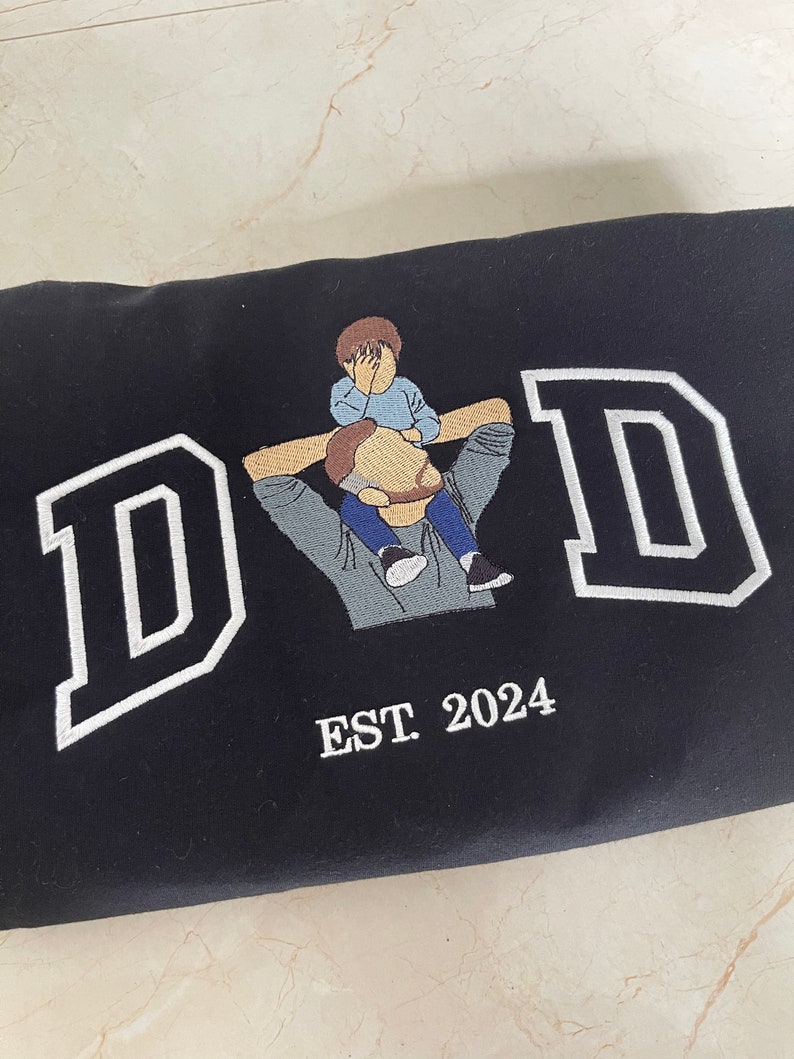 K&ouml;nnte beinhalten: Schwarzer Sweatshirt mit wei&szlig; gesticktem Text "DAD" und einem kleinen gestickten Bild von Vater und Sohn. Der Text "EST. 2024" ist unter dem Bild gestickt.