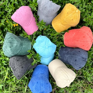 Pode incluir: Dez bon&eacute;s de beisebol de diferentes cores dispostos em c&iacute;rculo sobre um leito de grama verde. Os bon&eacute;s s&atilde;o feitos de um tecido macio e texturizado e v&ecirc;m em uma variedade de cores, incluindo azul, verde, vermelho, rosa, amarelo e cinza.