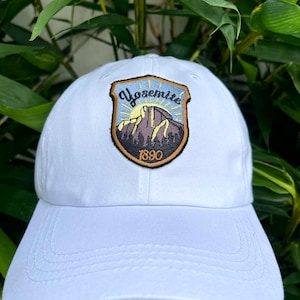 May include: White baseball cap with a brown and black embroidered patch featuring a mountain scene and the text "Yosemite 1890".