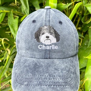 Pu&ograve; includere: Un cappello da baseball in denim blu scuro con un viso di cane bianco e nero ricamato e il nome "Charlie" cucito in bianco.