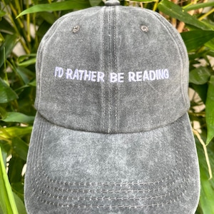 Peut inclure: Une casquette de baseball gris foncé avec le texte "I'd rather be reading" brodé en violet clair sur le devant.